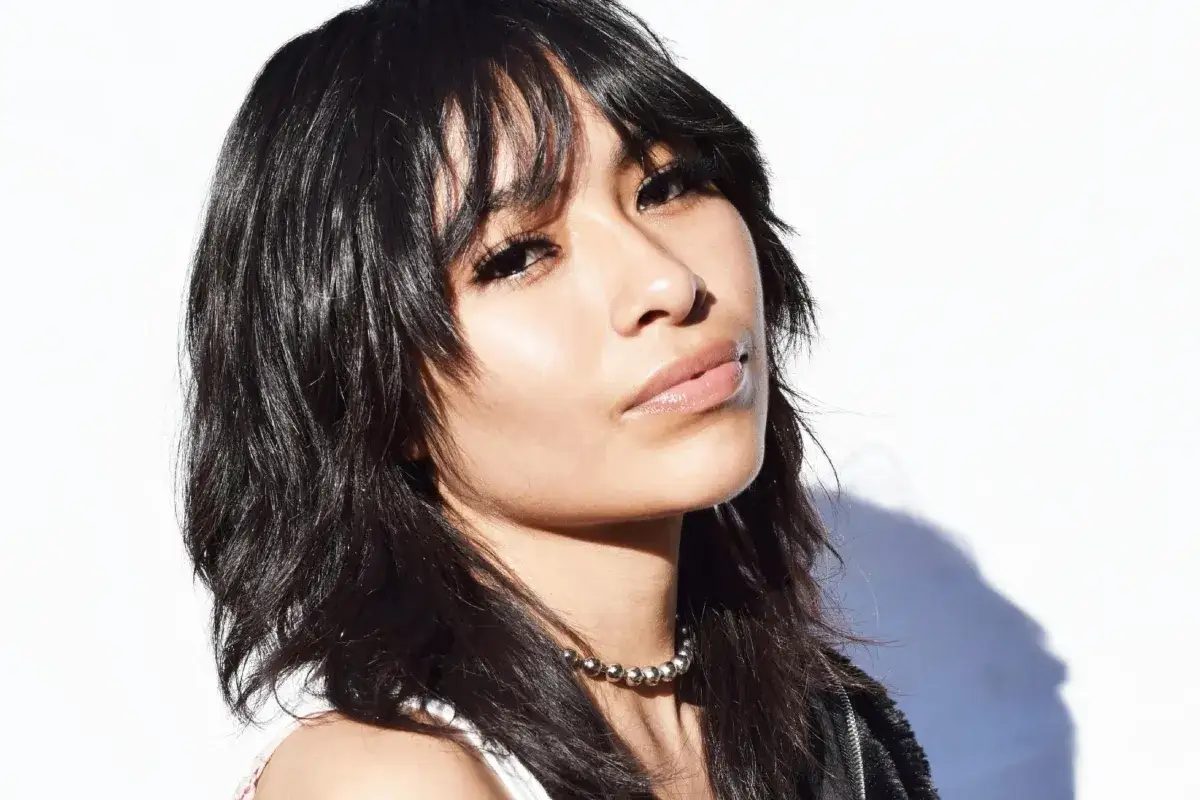 Joven con un moderno mullet corto, cabello oscuro y labios brillantes, luciendo un collar de cuentas plateadas.