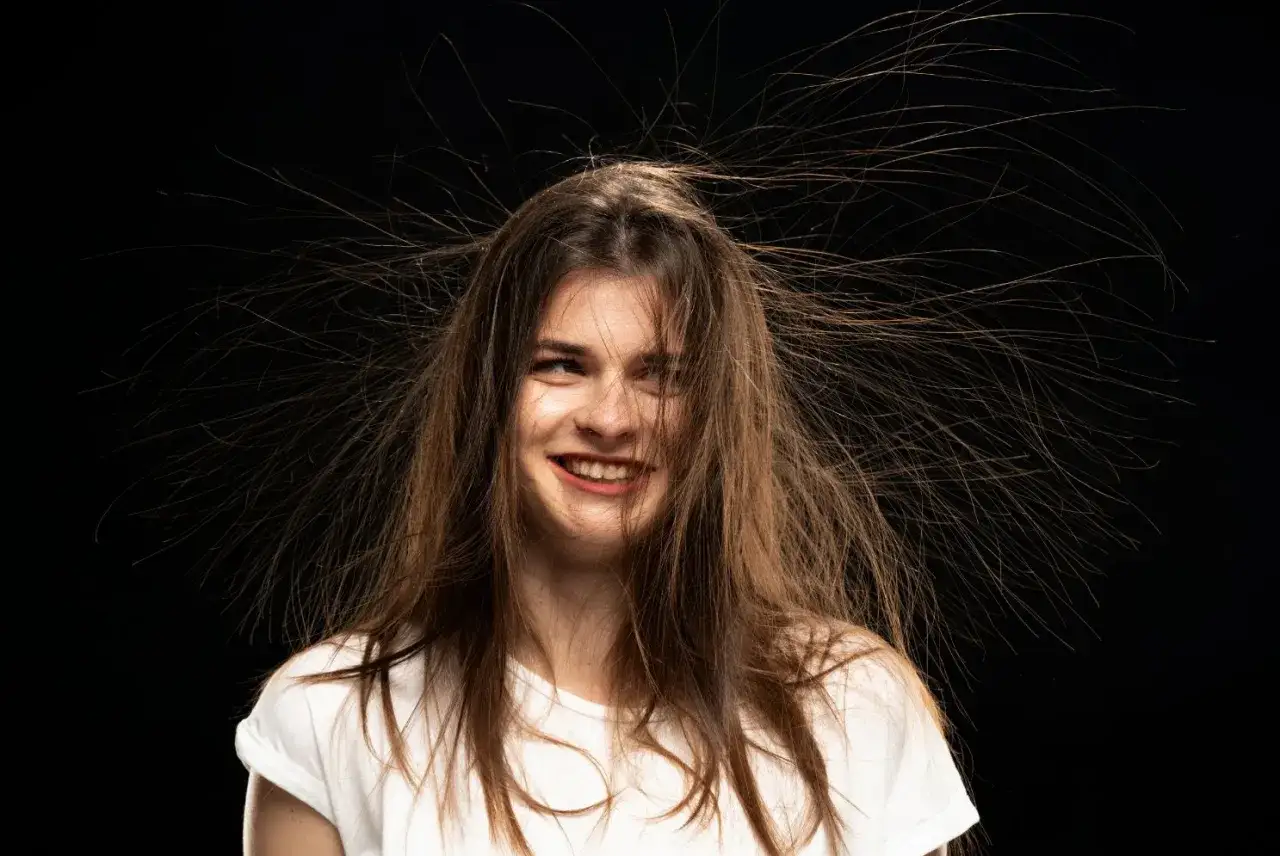 Mujer con pelo electrizado, como si buscara c&oacute;mo quitar la electricidad del pelo. Su cabello se alza en todas direcciones, creando un efecto c&oacute;mico.