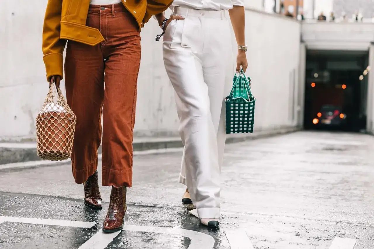 Dos mujeres caminan con estilo. Una lleva pantalones terracota y botas, la otra, pantalones blancos y un bolso verde. ¡Un look genial para quienes saben como ser bajita y lucir fabulosas!