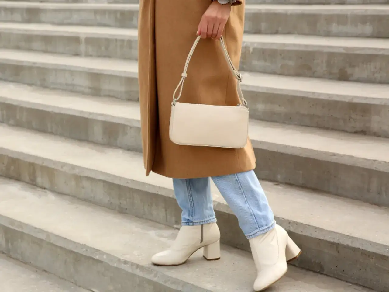 Mujer combinando botas beige con jeans y abrigo camel. Un bolso blanco completa el look.