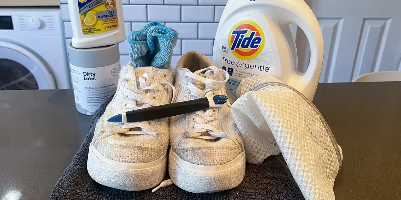 Un par de zapatillas blancas de tela sucias listas para limpiar con un cepillo, detergente Tide y un potenciador de lavandería.