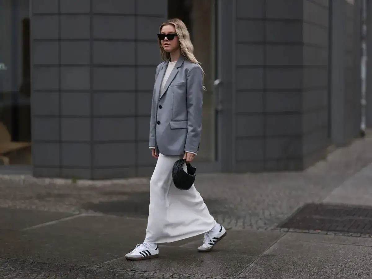 Mujer con zapatillas blancas y grises, falda larga blanca, blazer gris y bolso negro.