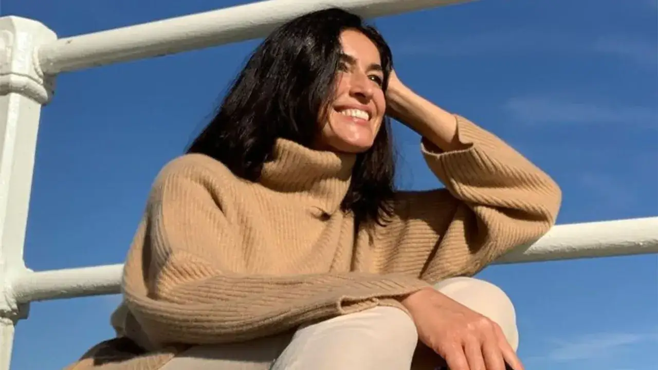 Blanca Romero sonríe con un jersey de cuello alto color camel, apoyada en una barandilla blanca bajo un cielo azul.