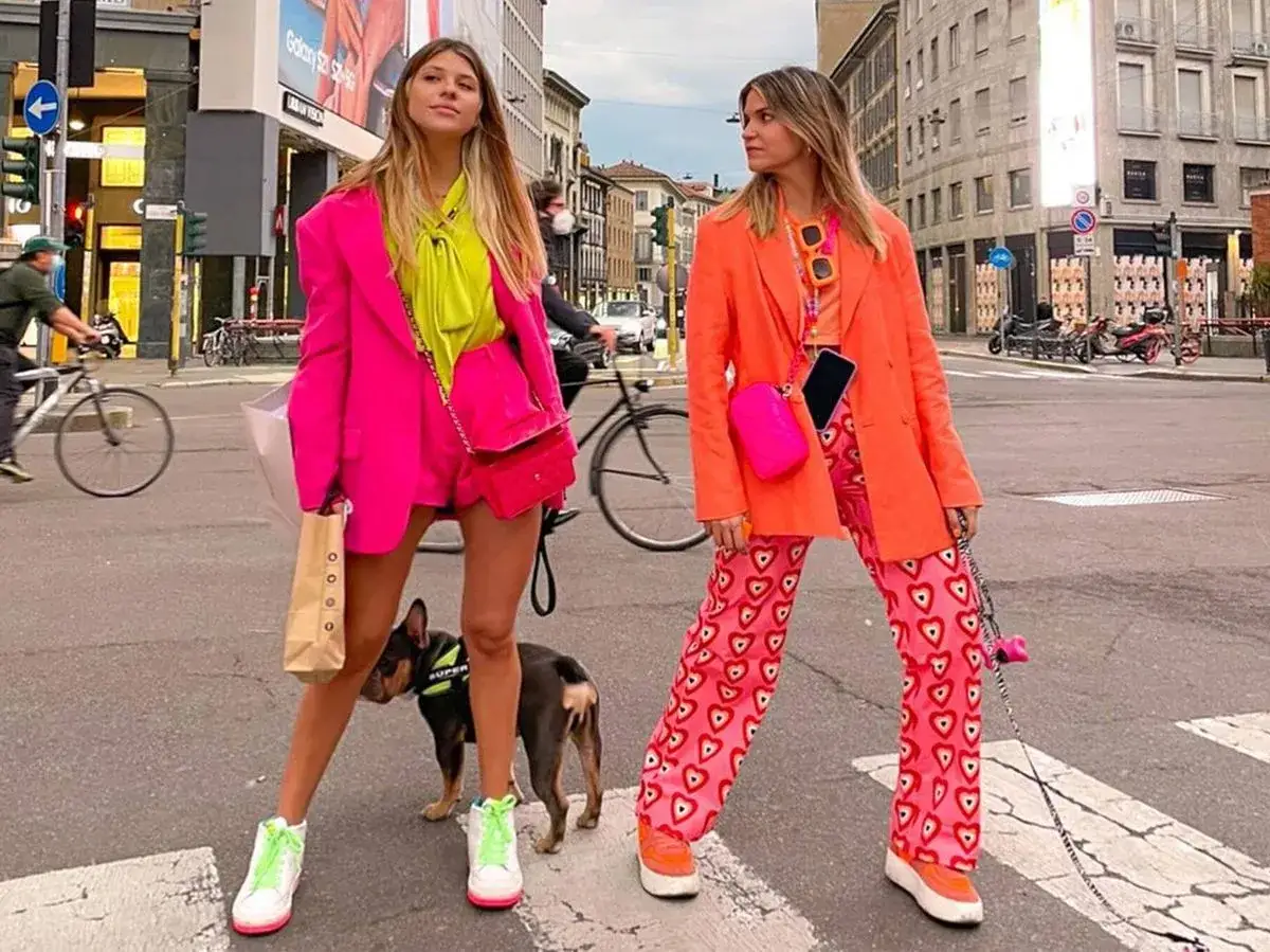 Dos mujeres con trajes vibrantes, uno fucsia y otro naranja, pasean a un perro. Los colores que combinan con amarillo son el rosa y el naranja, creando un look audaz.