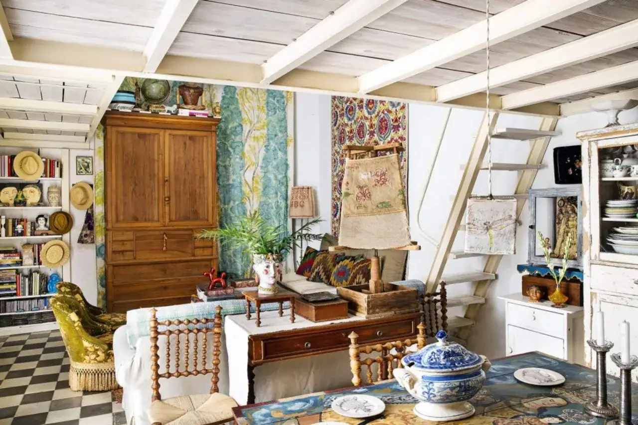 Un salón con estilo cottagecore, lleno de texturas, colores y objetos vintage. Un armario de madera, estanterías repletas y una escalera rústica crean un ambiente acogedor.