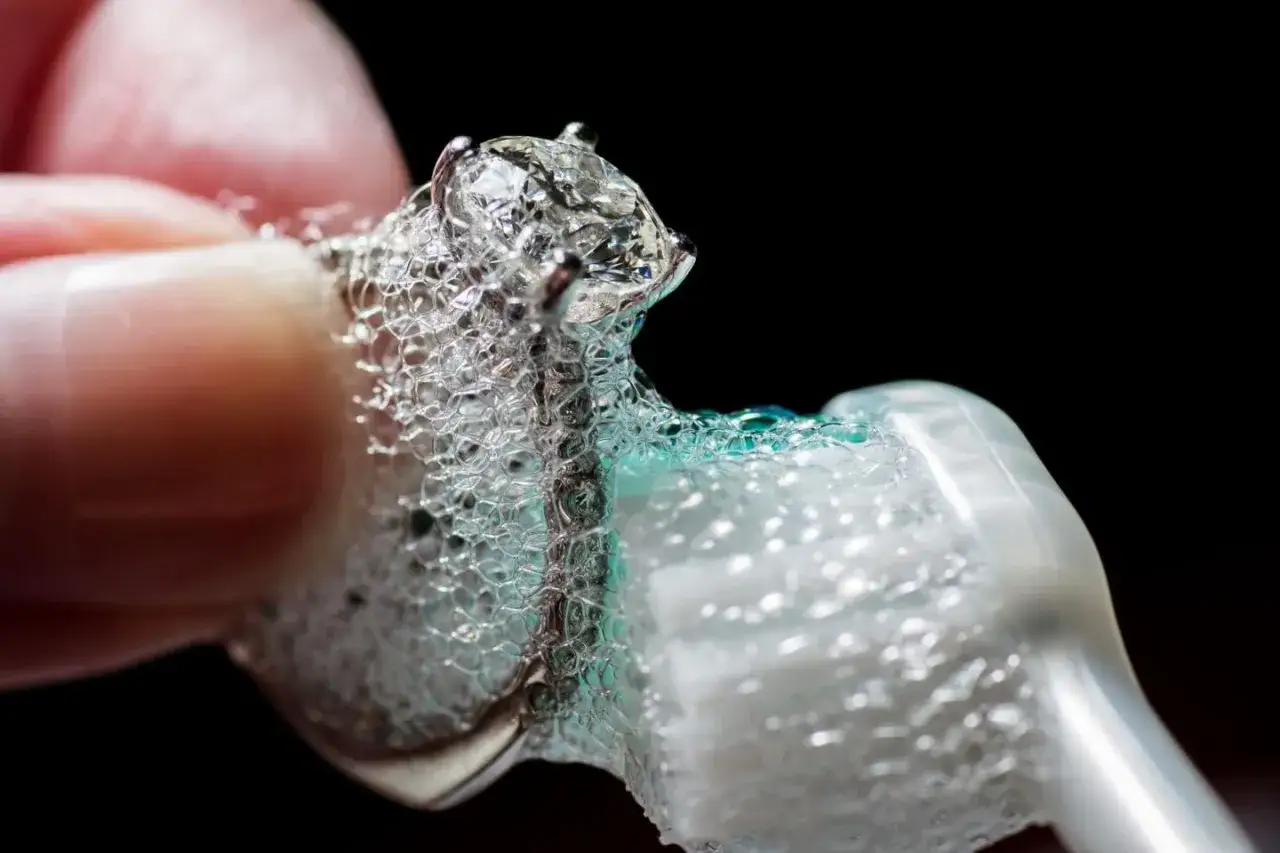 Un dedo sostiene un anillo con diamantes mientras una cepillo de dientes con espuma lo ayuda a limpiar joyas.