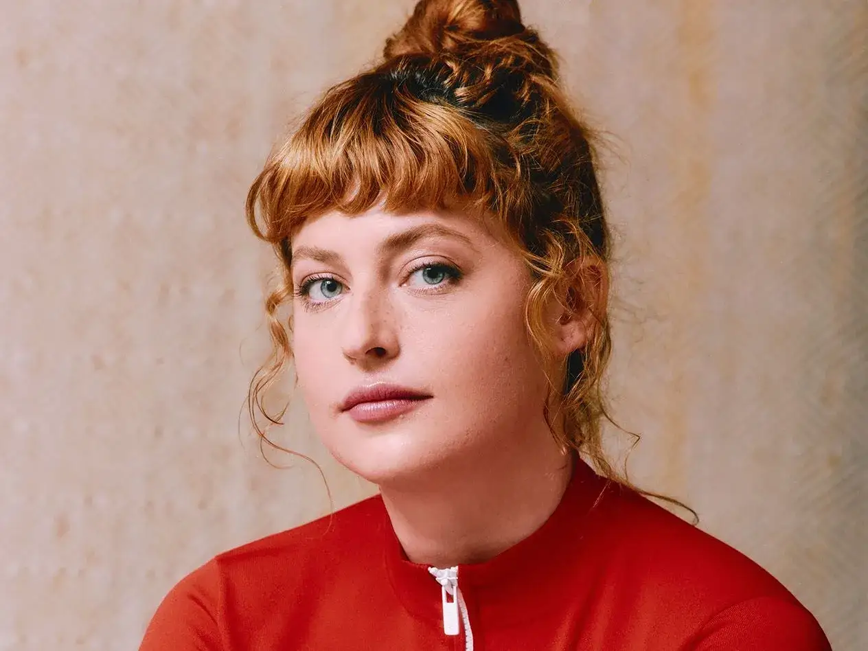 Chloe Wallace, con su cabello rojizo recogido en un moño y flequillo despeinado, mira fijamente con sus ojos azules.
