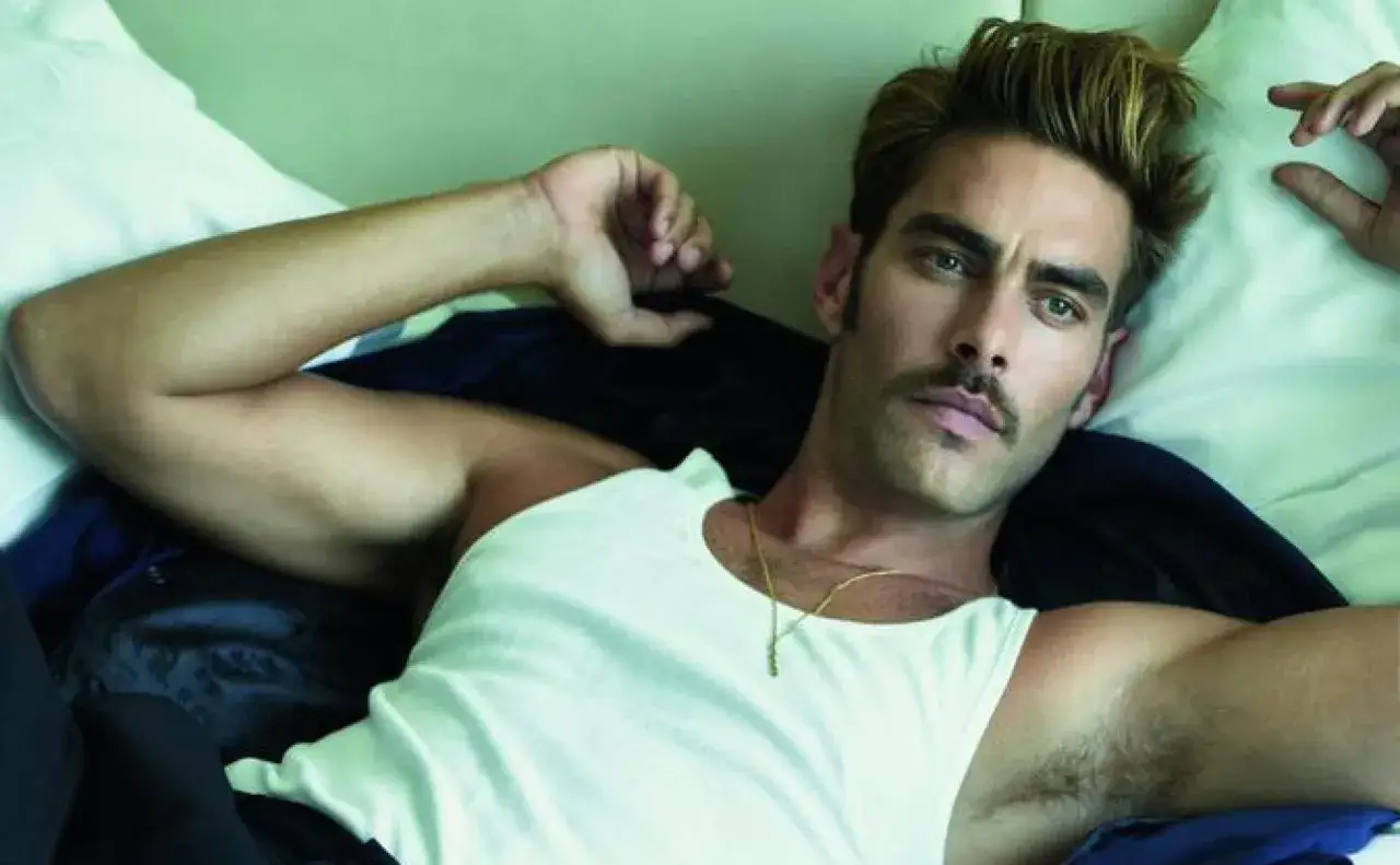 Jon Kortajarena, modelo español, recostado en una cama, con un bigote y un collar dorado.