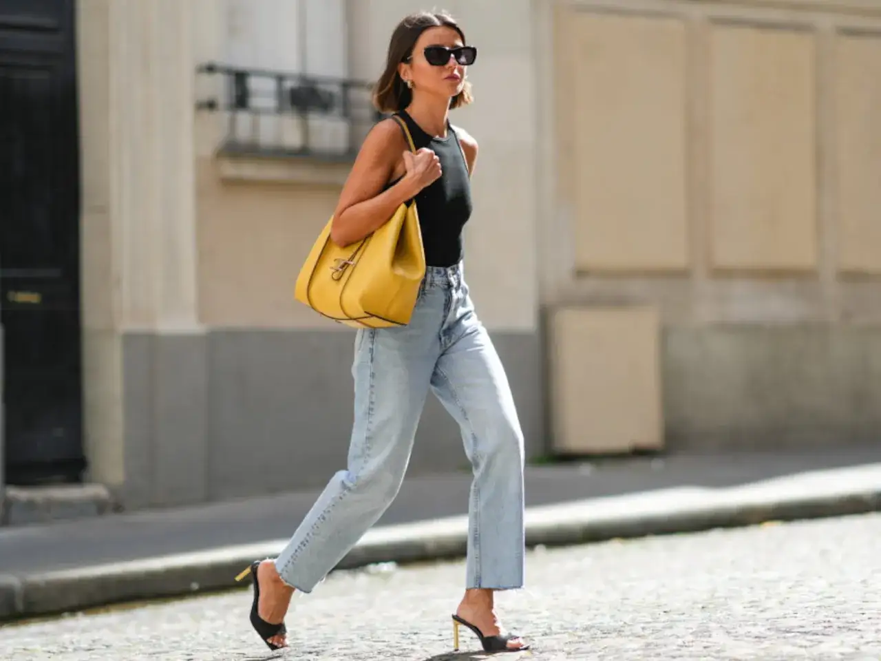 Mujer con vaqueros anchos y top negro, bolso amarillo y sandalias de tacón. Su figura estilizada, con sutiles hip dips, se mueve con elegancia por la calle.