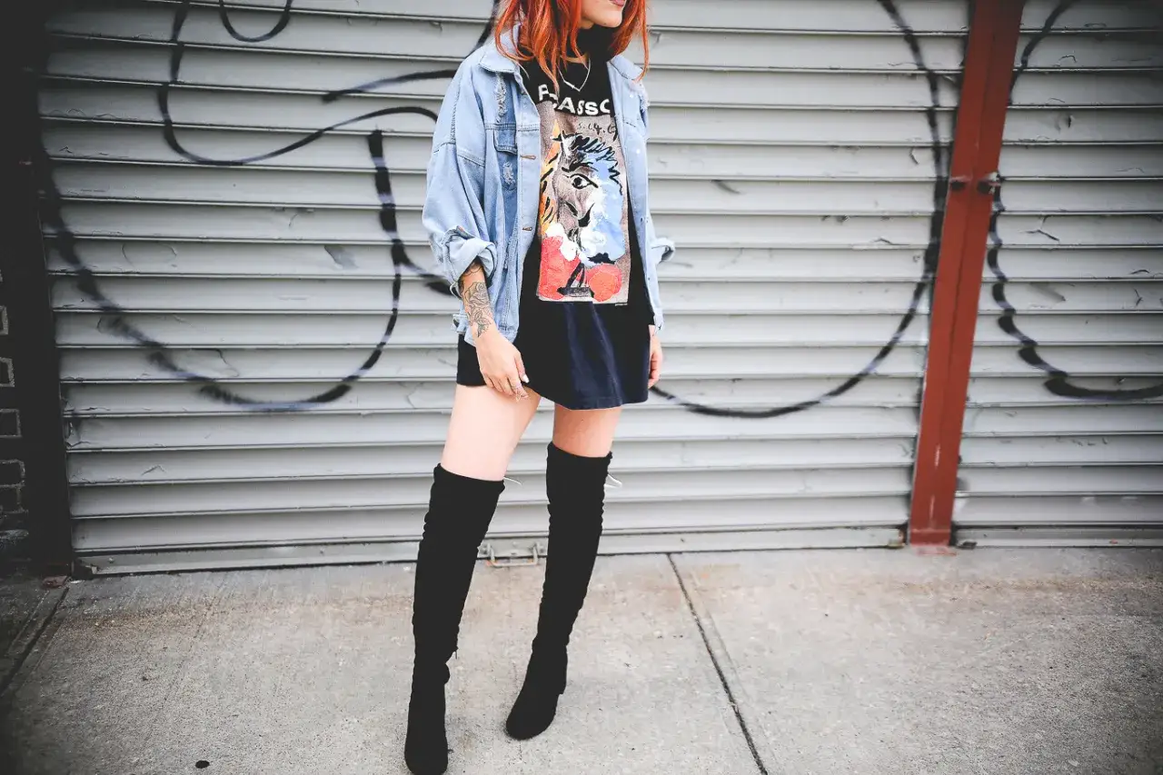 Chica con botas altas para bajitas, chaqueta vaquera y camiseta gráfica, posando frente a una pared de garaje con grafitis.