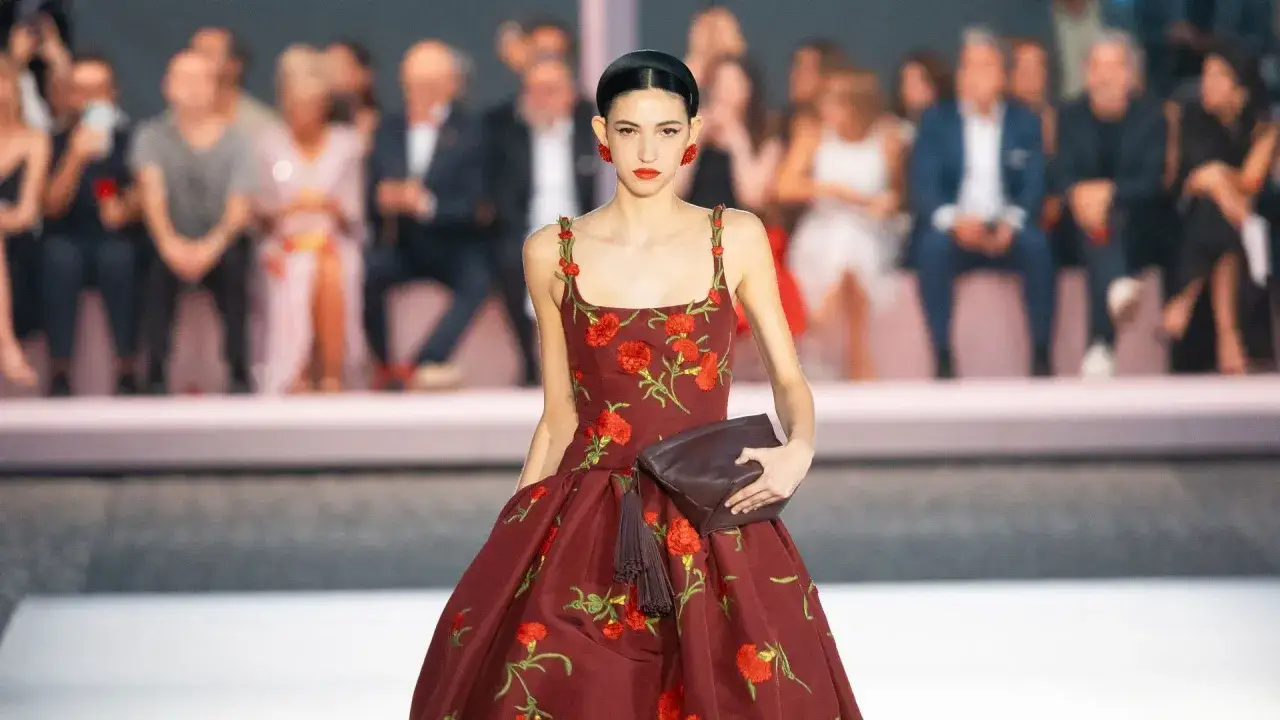 Una modelo luce un vestido rojo con flores rojas, un bolso marrón y pendientes rojos. La pregunta de quién es Carolina Herrera se responde con este elegante diseño.
