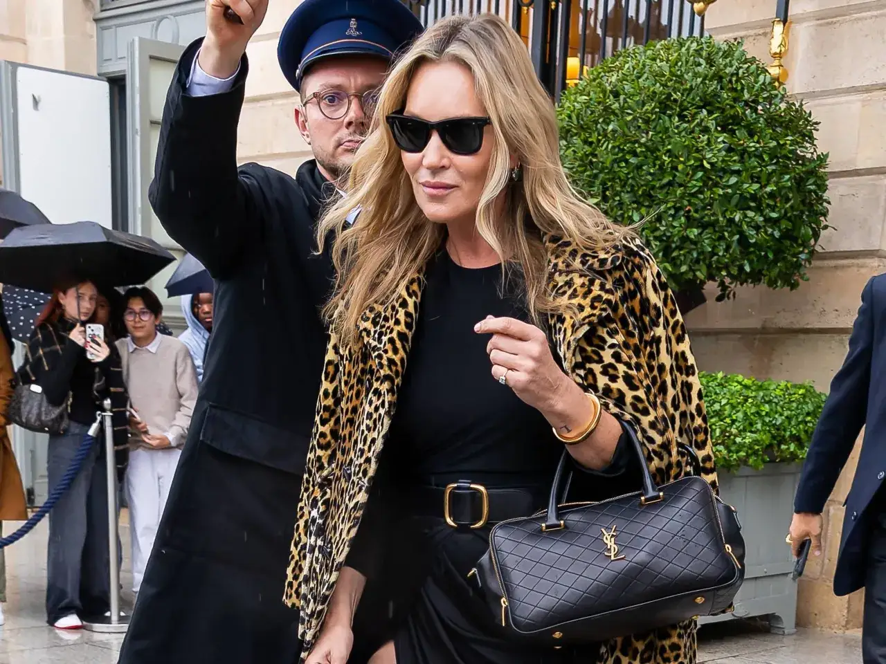 Kate Moss, con su icónico abrigo de leopardo y gafas de sol, camina con elegancia. Un guardaespaldas la acompaña.