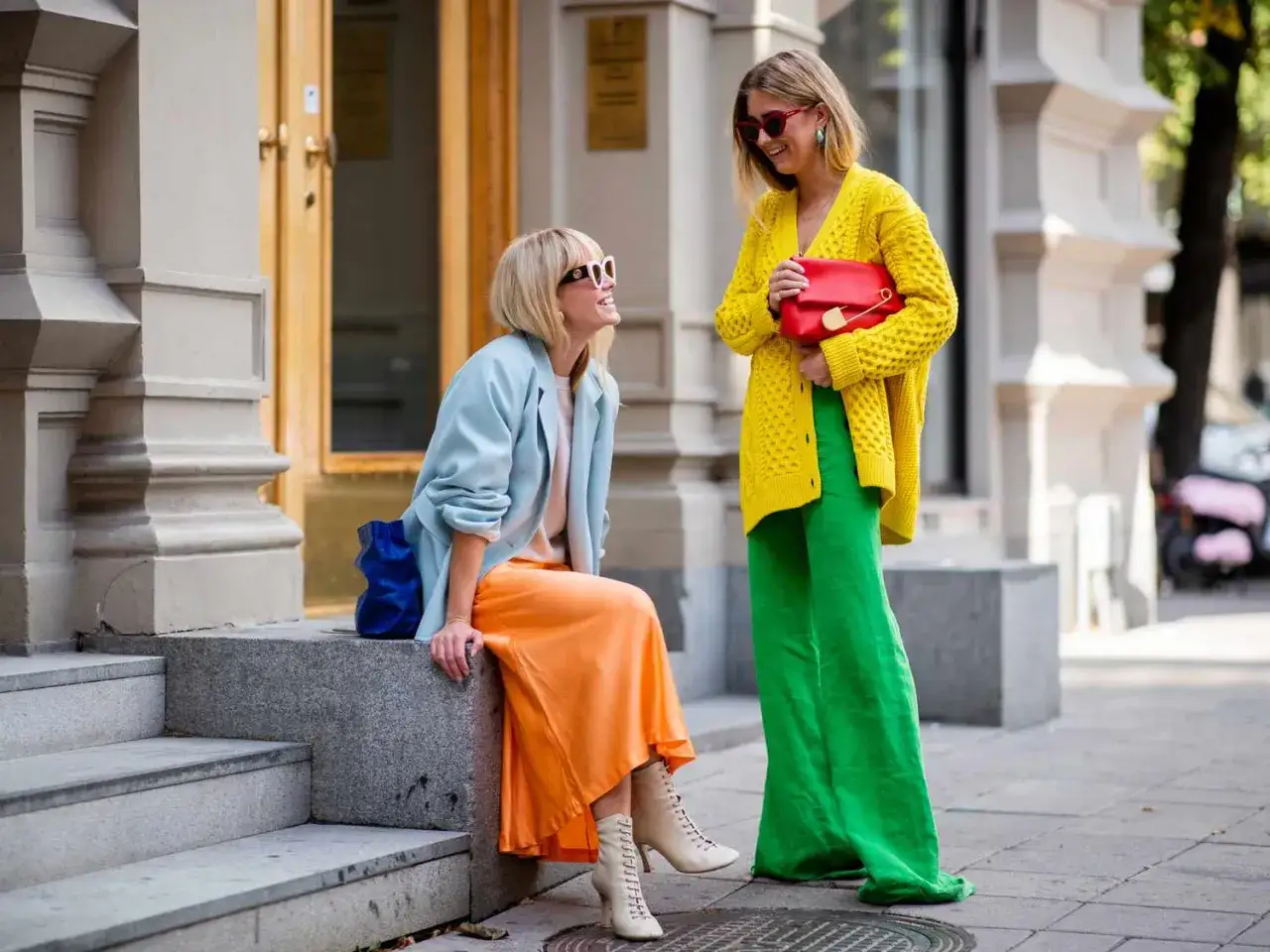 Dos mujeres lucen atuendos vibrantes de color block: una con chaqueta azul y falda naranja, la otra con cárdigan amarillo y pantalones verdes.