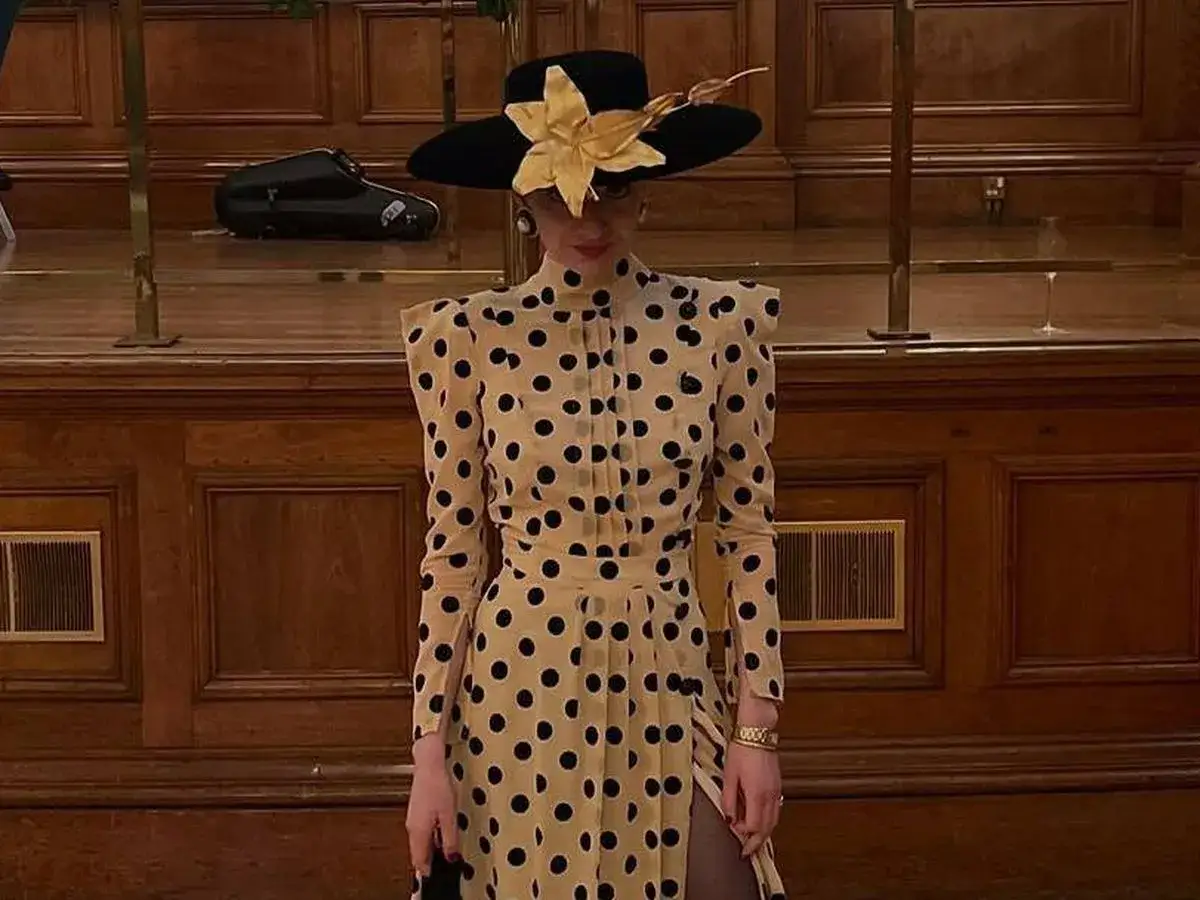 Blanca Llandres, con un elegante vestido de lunares y un sombrero negro adornado, posa ante un fondo de madera.