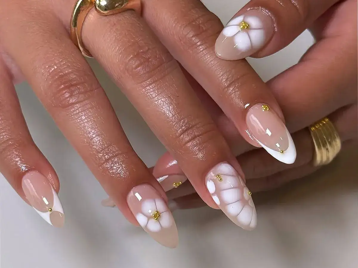 Manicura con diseño de flores blancas y detalles dorados. Uñas almendradas con base nude y francesa blanca, adornadas con pequeñas flores blancas y puntos dorados.