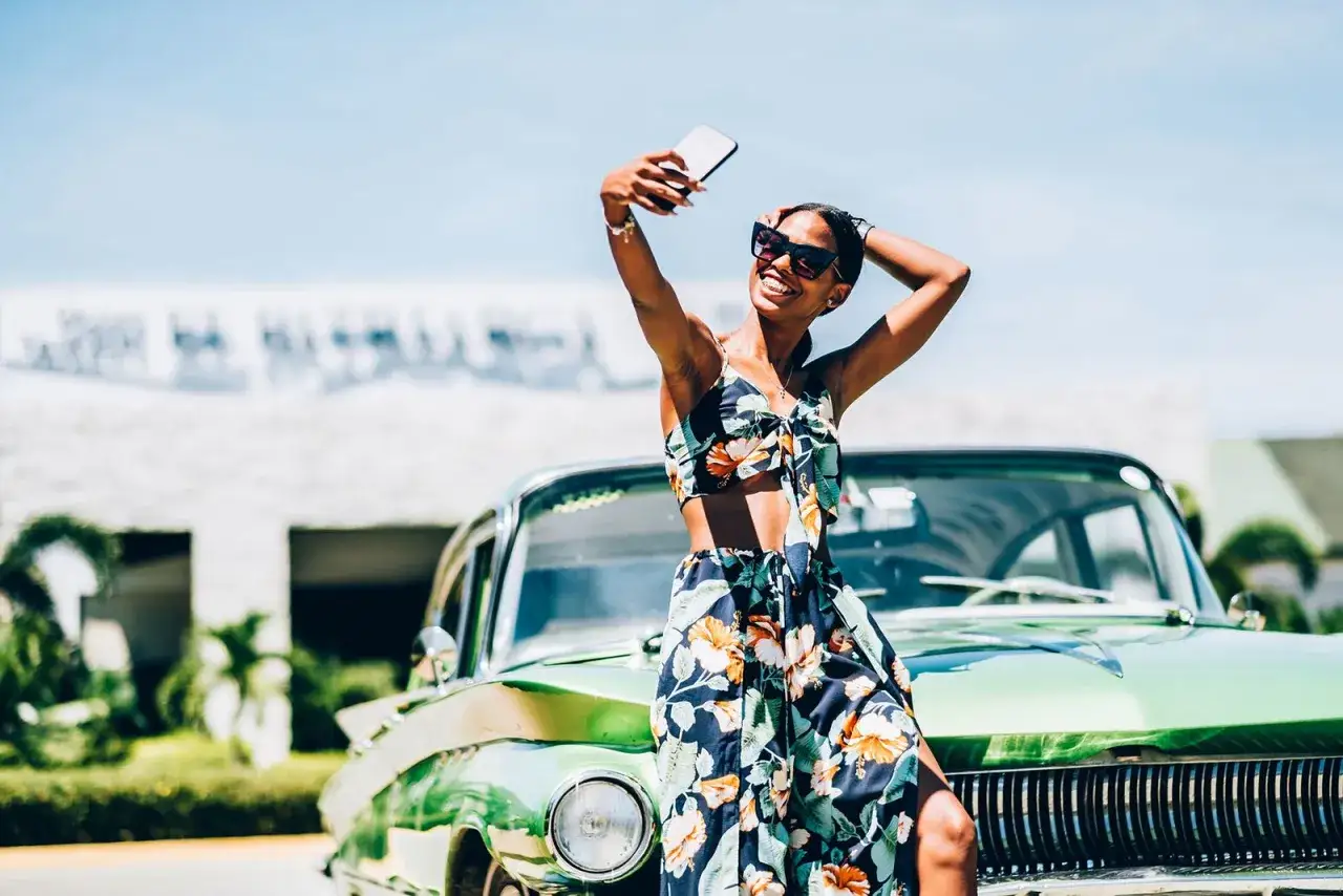 Chica vsco sonriendo y tomándose una selfie junto a un coche clásico verde bajo el sol.