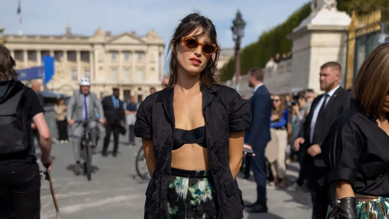 Mujer con gafas de sol y top negro, luciendo un estilo chic que evoca las marcas francesas. Al fondo, un edificio histórico y gente paseando.