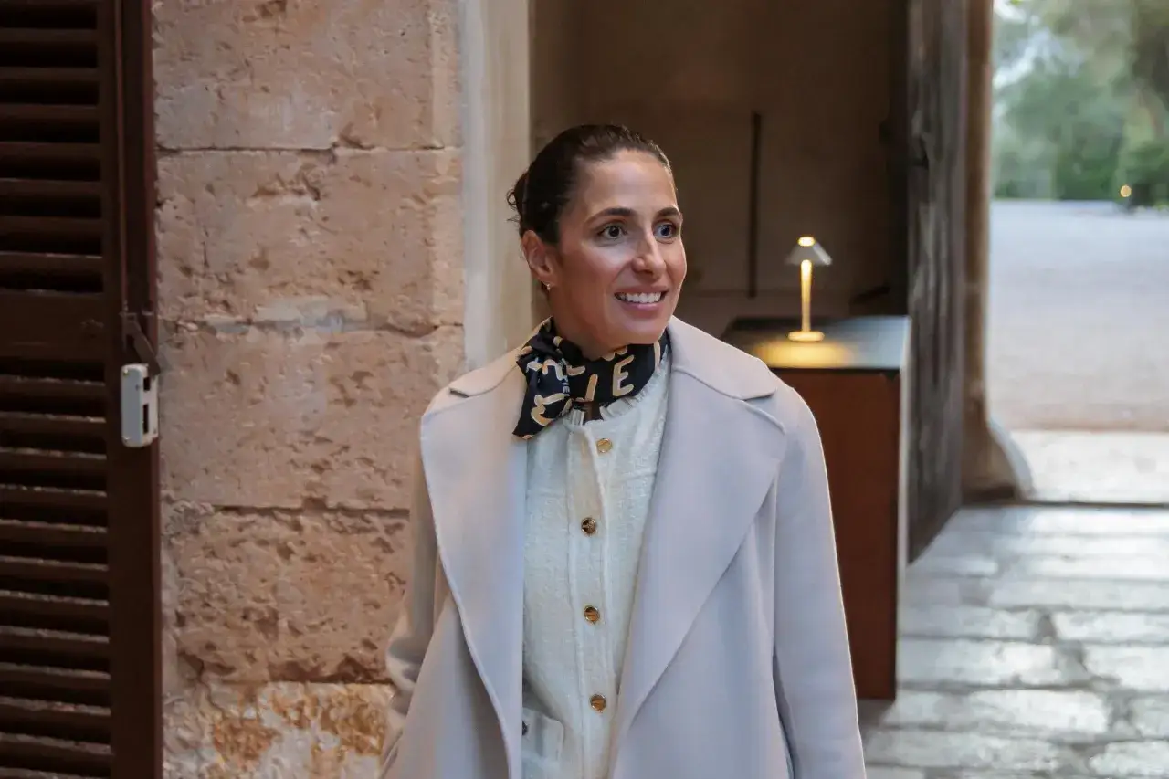 Xisca Perelló, sonriente y elegante con abrigo claro y pañuelo estampado, camina por un patio de piedra.