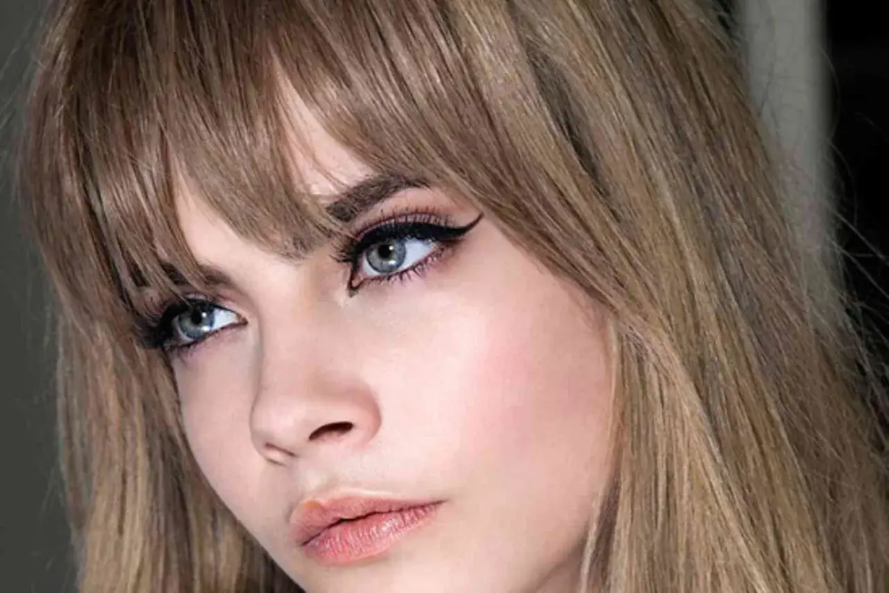 Primer plano de una mujer con flequillo rubio desfilado, delineado de ojos dram&aacute;tico y labios rosados.