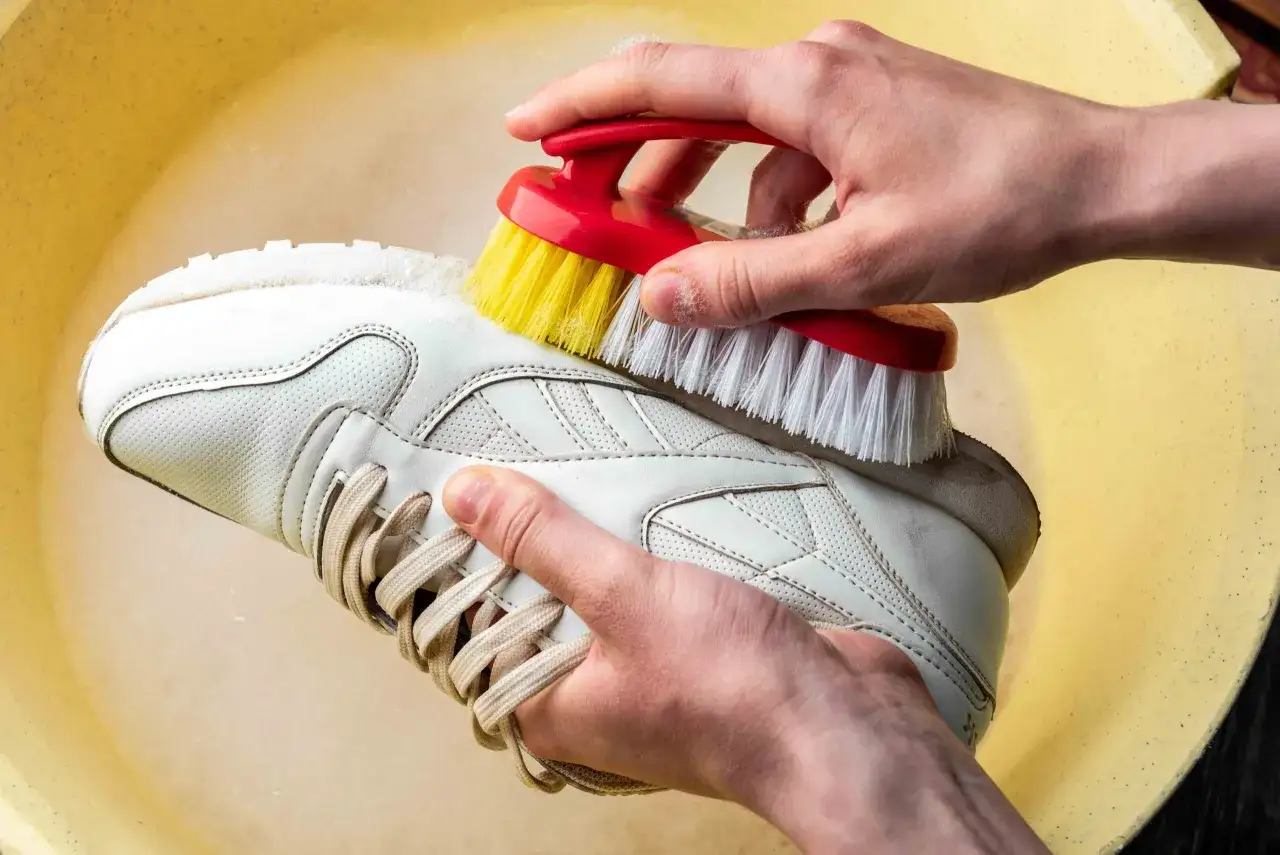 Manos limpiando un zapato de piel blanco con un cepillo rojo y amarillo en una tina amarilla.