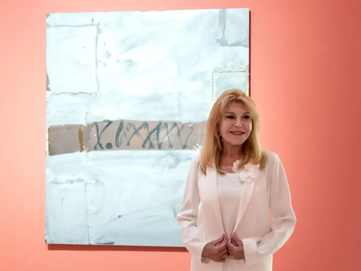 Tita Thyssen posa sonriente junto a un cuadro abstracto de tonos blancos y marrones.