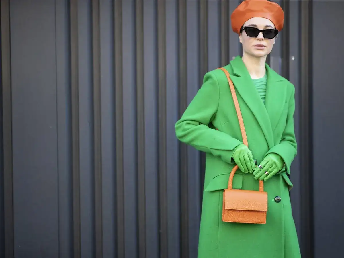 Mujer con abrigo verde vibrante, guantes a juego y un bolso naranja. Su boina y gafas de sol completan el look.