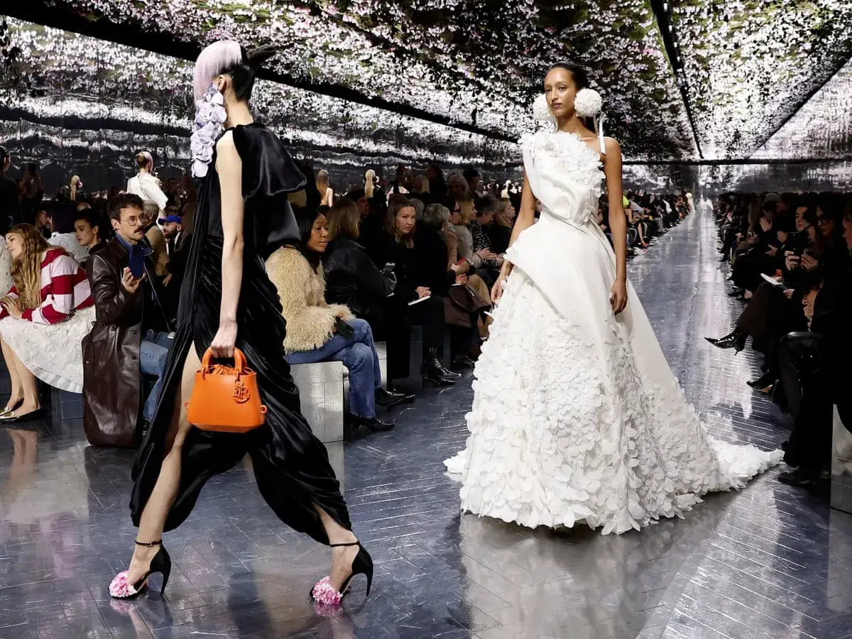 Modelos desfilan en la pasarela de Jonathan Anderson, una con vestido negro y bolso naranja, otra con un voluminoso vestido blanco.